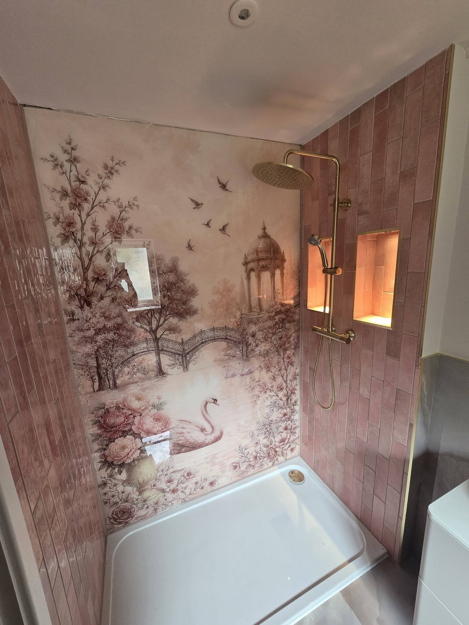 A shower wall with a pink floral mural panel, gold shower and a lit niche