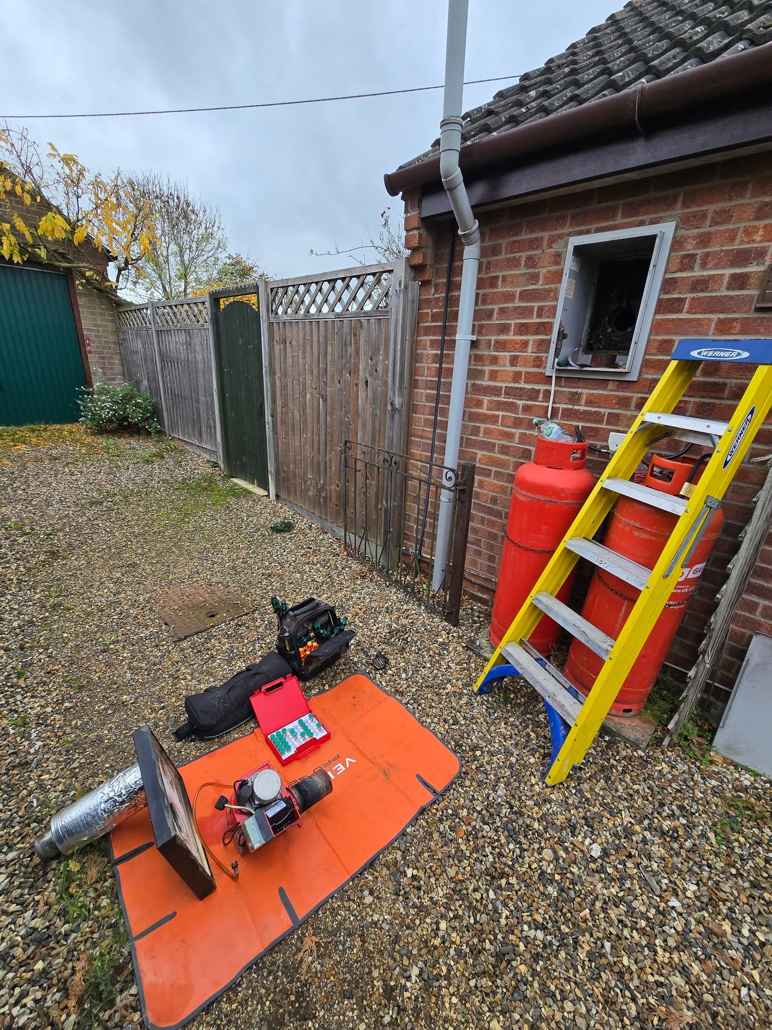 Tools and a boiler burner laid out on an orange mat beside a bungalow